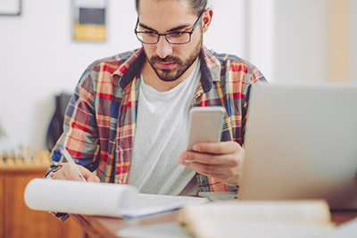 A photo of a young man holding a cell phone and looking at a pad of paper