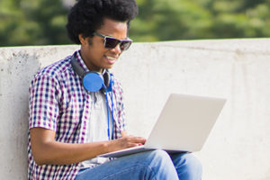 An image of a student sitting in the sun and working on his laptop.