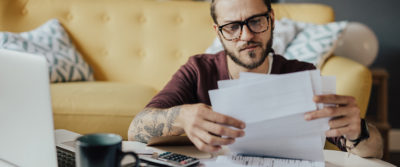 An image of a young man looking over paperwork in front of his laptop computer.